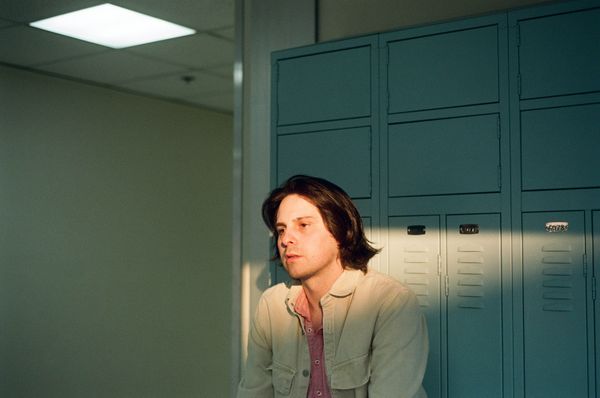 The artist EROCY seated in front of blue lockers with a faint strip of sunset light streaking across his face.